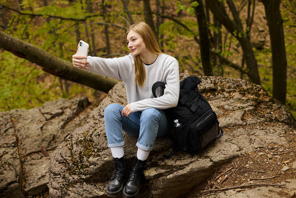 blonde young woman taking selfie sitting on rock at halt in the forest while hiking - Φωτογραφία, εικόνα