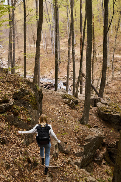 Back view of young blonde female traveler wearing backpack running to forest stream - Foto, immagini