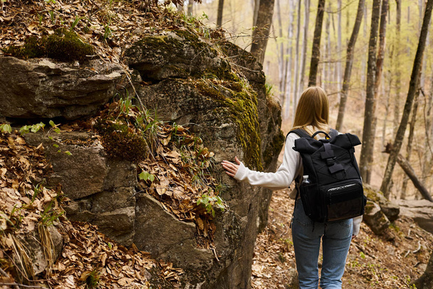 Back view of blonde female traveler wearing backpack on hiking trip and walking near rocky cliff - Foto, afbeelding