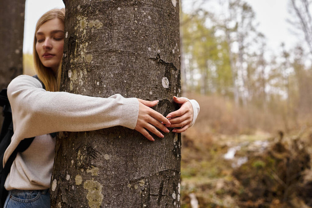 Jeune randonneuse blonde détendue avec sac à dos de voyage, étreignant un arbre et profitant d'un voyage paisible - Photo, image