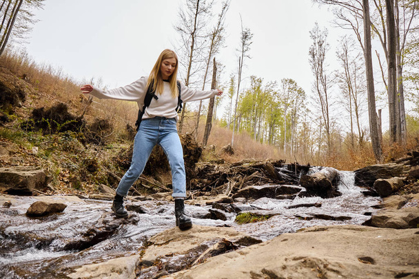 Joyful blonde female hiker wearing sweater and jeans crossing the forest creek jumping over water - Photo, Image