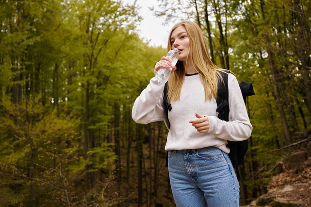 Podróżniczka w swetrze i dżinsach przemierzająca potok leśny spacerując po lesie pijąc wodę - Zdjęcie, obraz