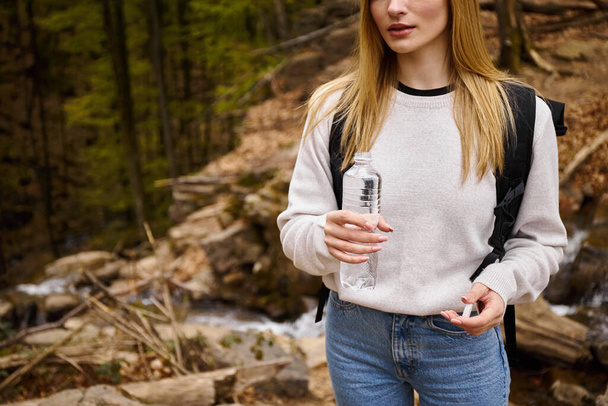 Woman traveler wearing sweater and jeans crossing the forest holding bottle of water - Photo, Image