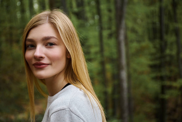 Uśmiechnięta, wesoła, piękna blondynka patrząca w kamerę podczas spaceru po lesie - Zdjęcie, obraz