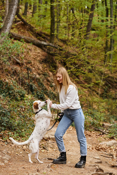 Χαρούμενη γυναίκα εκπαίδευση κατοικίδιο ζώο σκυλί της κρατώντας λουρί σε πεζοπορία υπόλοιπο με θέα στο βουνό και το δάσος - Φωτογραφία, εικόνα