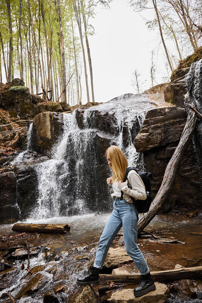 Active blonde female hiker crossing the forest creek walking on rocks near waterfall - Photo, Image