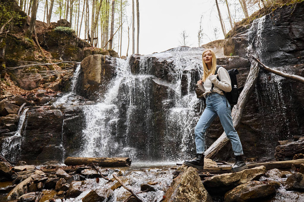 Young blonde female hiker crossing the forest creek walking on rocks near waterfall - Foto, afbeelding
