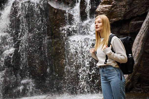 Porträt einer jungen blonden Reisenden, die in einem wunderschönen Wald wandert und in der Nähe eines Wasserfalls steht - Foto, Bild