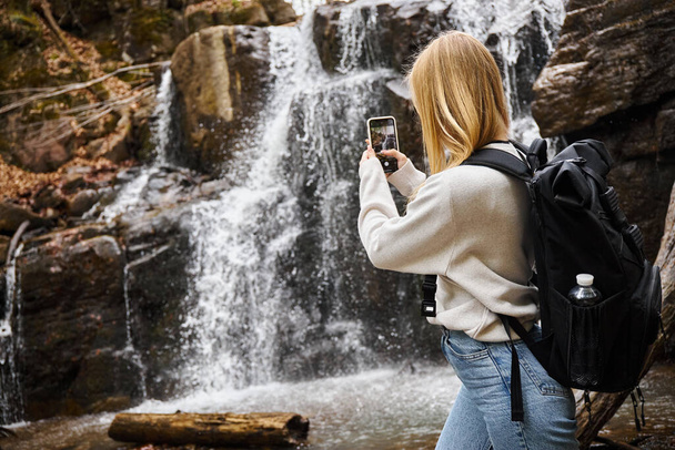 Rückansicht der blonden jungen Frau, die beim Wandern einen Wasserfall im Wald fotografiert - Foto, Bild