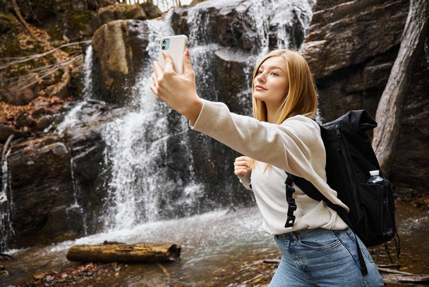 Blondynka młoda kobieta robi selfie w pobliżu górskiego wodospadu w lesie podczas wędrówki - Zdjęcie, obraz