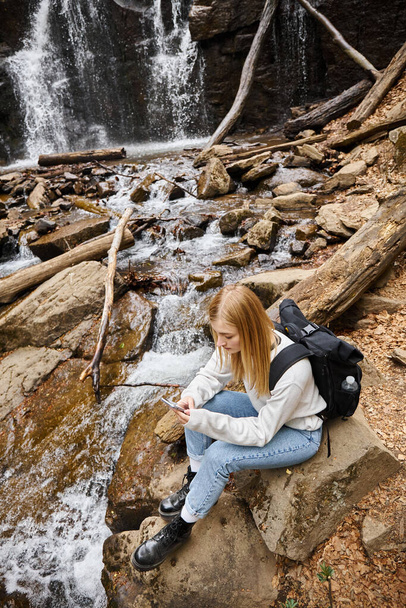viaggiatore donna bionda utilizzando il telefono seduto vicino alla cascata di montagna nella foresta durante le escursioni - Foto, immagini
