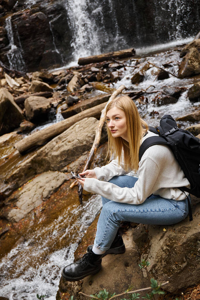 blondynka trzyma telefon siedzi w pobliżu górskiego wodospadu w lesie podczas wędrówki - Zdjęcie, obraz