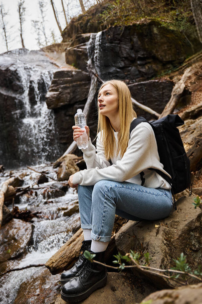 Young woman with backpack sitting and holding bottle of water and looking at forest near waterfall - 写真・画像