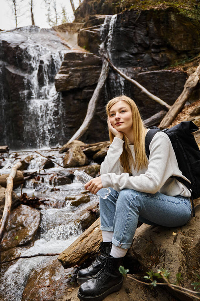 Mujer sonriente sentada con botella de agua y mirando hacia otro lado en el bosque cerca de la cascada - Foto, Imagen