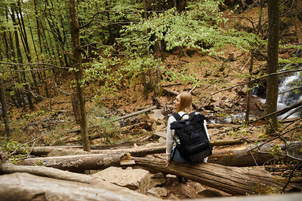 Vista posterior de la joven senderista rubia con mochila negra sentada y descansando durante el trekking - Foto, Imagen