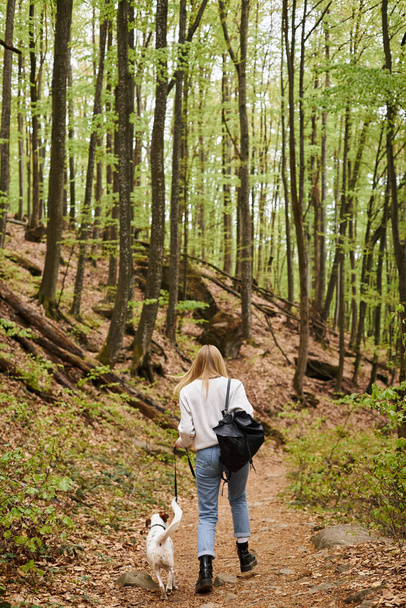 Back view of blonde young active woman walking her dog companion at forest backpacking trip - Foto, imagen