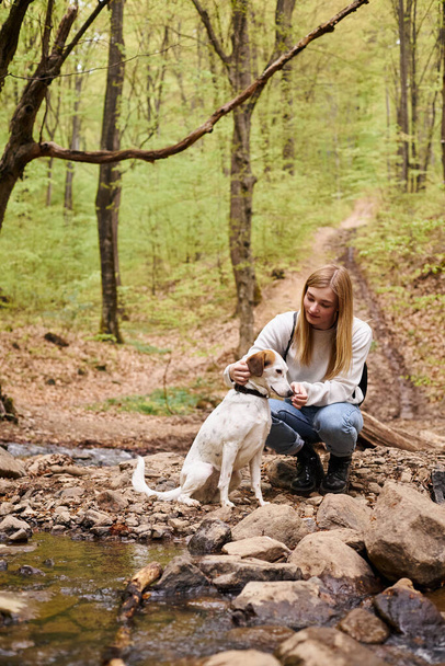 Smiling woman hugging gently her dog companion and looking at pet while walking in forest - Photo, Image