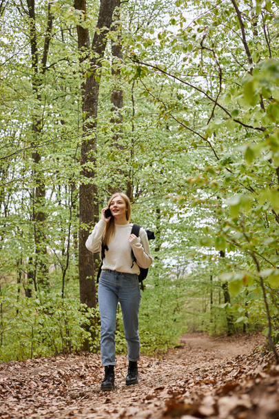 Pretty blonde woman traveler with backpack talking by phone walking in forest scenery - Photo, Image