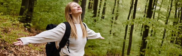 Sonriente mujer rubia relajada excursionista con suéter y mochila con los brazos abiertos en el bosque, pancarta - Foto, Imagen