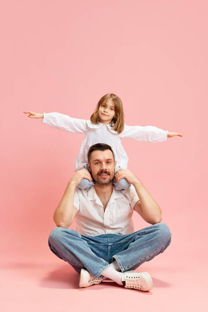 Homem sorridente sentado de pernas cruzadas com a menina nos ombros, que estendem os braços como avião contra fundo pastel rosa. Conceito do Dia Internacional da Felicidade, infância e paternidade, emoções. Anúncio - Foto, Imagem