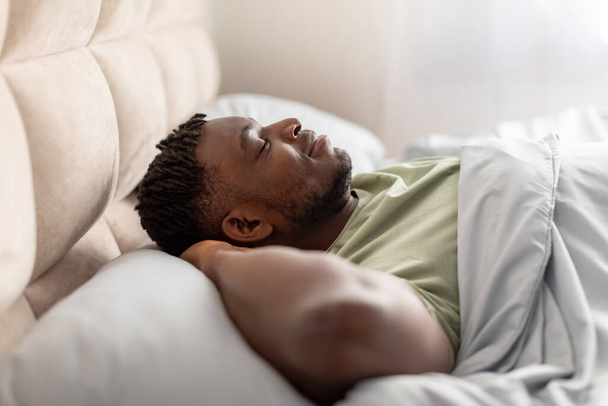 Oldalnézet a békés fekete Y-generációs férfi mélyen alszik borított fehér takaró, kéz a fej mögött nyugodt pózban, miközben szunyókál a hazai hálószoba belső. Az egészséges pihenés fogalma - Fotó, kép