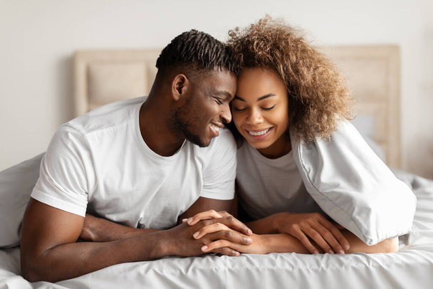 Szczęśliwa para czarnych kochanków w łóżku trzymająca się za ręce i dotykająca czoła, flirtująca rano, ciesząca się intymnością i romantyzmem w swojej sypialni w domu. Małżeństwo i związek - Zdjęcie, obraz
