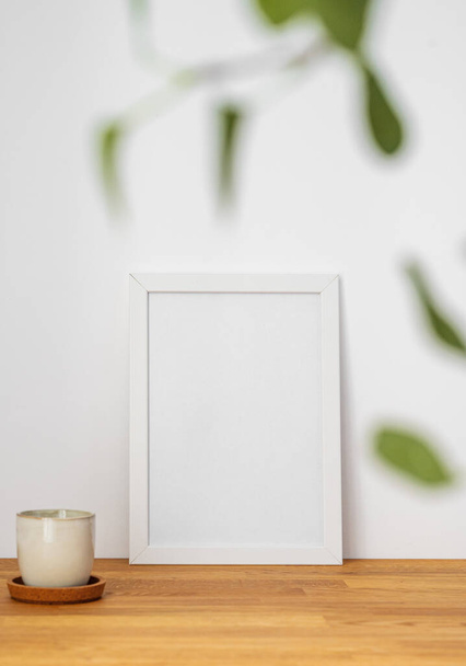 Bílý rám s prázdným plátnem opřeným o světlou stěnu a na dřevěném stolku s rozmazaným zeleným listím a šálkem čaje. Dispoziční koncept pro fotografii, plakát nebo malbu. Kopírovat prostor. - Fotografie, Obrázek