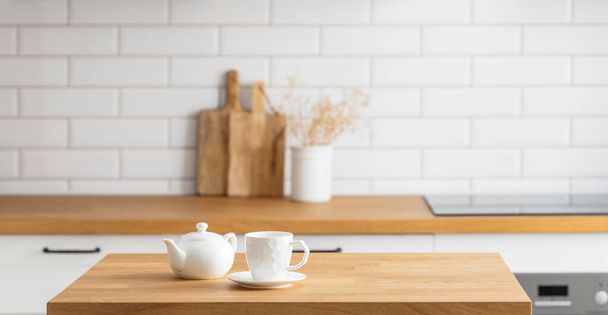 Wooden oak table with a cup of tea and a kettle in front of the kitchen with a white brick background with cutting boards and a vase with dry branches. Cozy breakfast concept in Scandinavian style.  - Photo, Image