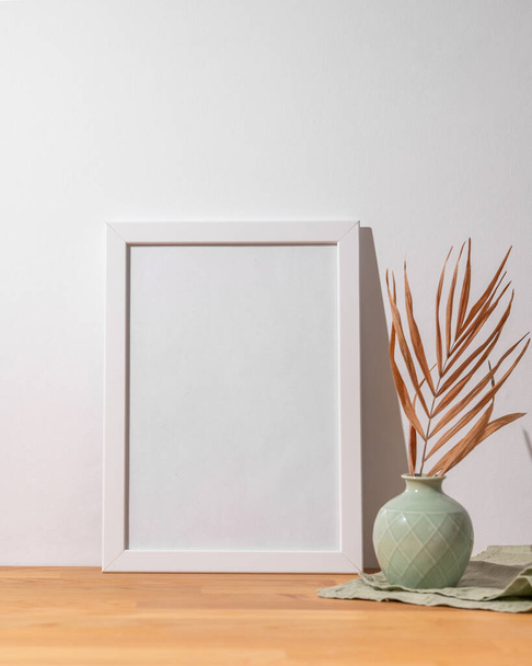 Bílý rám s prázdným plátnem opřeným o světlou stěnu a na dřevěném stolku s vázou. Dispoziční koncept pro fotografii, plakát nebo malbu. Kopírovat prostor. - Fotografie, Obrázek