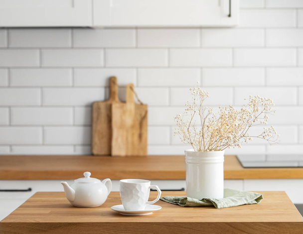 Wooden oak table with a cup of tea, a kettle and dry flowers in a vase  in front of the kitchen with a white brick background. Cozy breakfast concept in Scandinavian style.  - Photo, Image