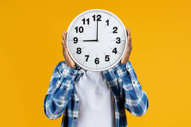 Unrecognizable black male holding large round clock in front of his face, african american man in plaid shirt standing on yellow background, representing concept of time management and punctuality - Photo, Image