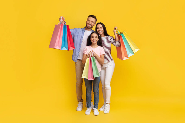 Концепция продаж. Счастливая кавказская семья из трех человек, держащих в руках сумки с новой одеждой и подарками, счастливые родители и дочь-подросток, несущие покупки, позирующие на желтом фоне студии, полной длины - Фото, изображение