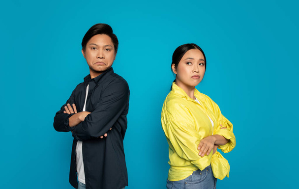 Stern-faced angry sad chinese man and woman with arms crossed, showing a defiant or confrontational attitude, standing against a solid blue background, studio. Relationships problems - Photo, Image