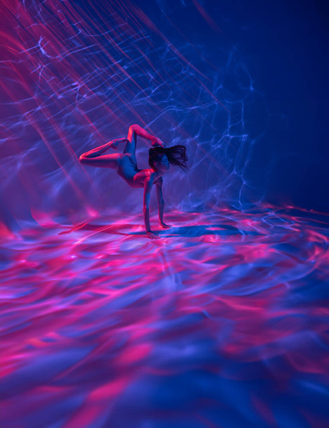 Unterwasser-Shooting der schönen Ballerina im Body, die durch rosa und rote Lichtstrahlen schwimmt und im Wasser tanzt. Ballerina vor Wasserhintergrund. - Foto, Bild