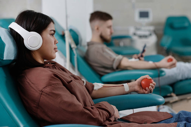 Женщина, слушающая музыку, сидящую в кресле во время переливания, мужчина, сдающий кровь на заднем плане - Фото, изображение
