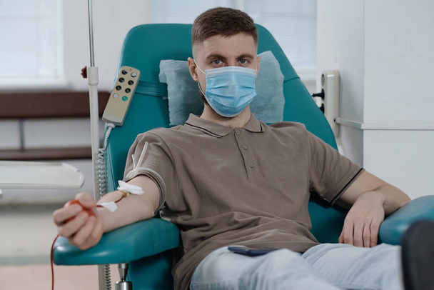Fiatal fehér férfi védő maszkban ül orvosi székben a vérátömlesztési folyamat alatt - Fotó, kép