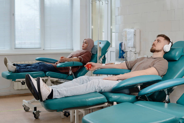 Hombre caucásico y mujer musulmana relajándose lado a lado en la clínica durante la donación de sangre - Foto, Imagen