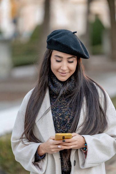 Mujer feliz en una boina negra usando su teléfono inteligente al aire libre con una sonrisa suave.  - Foto, Imagen