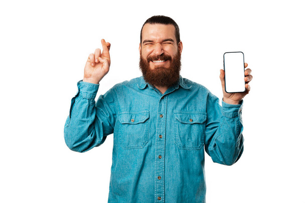 Позитивний бородатий чоловік тримає пальці схрещеними, тримаючи телефон з порожнім екраном. - Фото, зображення