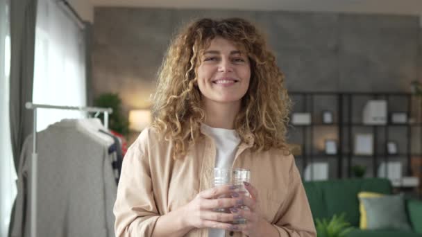 una donna bella femmina adulta a casa con un bicchiere d'acqua capelli ricci sorriso felice stile di vita sano concetto di idratazione - Filmati, video