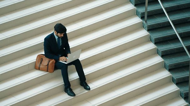 Widok inwestora pracującego lub planującego strategię przy użyciu laptopa na schodach. Profesjonalny biznesmen noszący garnitur podczas pracy i wpisujący analizę danych za pomocą laptopa w nowoczesnym hotelu. Ekstrakt. - Zdjęcie, obraz