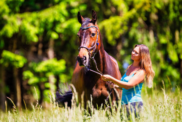 Een jonge vrouw ruiter met lang brunette haar staat met haar paard op een hoge zomerweide in de felle zon. - Foto, afbeelding
