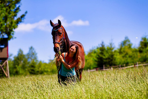 Een jonge vrouw ruiter met lang brunette haar staat met haar paard op een hoge zomerweide in de felle zon. - Foto, afbeelding