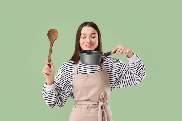 Mooie jonge vrouw in schort met kookpot en houten lepel op groene achtergrond - Foto, afbeelding