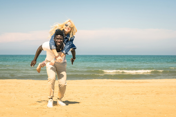 幸せな混血のボーイ フレンドとガール フレンドの愛の物語 - 人種差別多民族統合の愛の先頭に再生、ピギーバック ジャンプ - を楽しんでビーチで若い混血カップル - 写真・画像