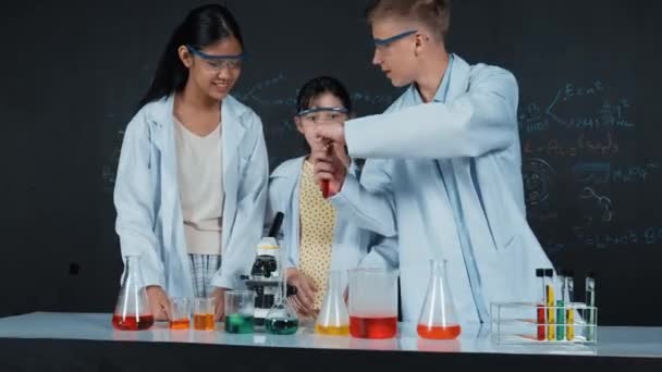 Smart boy mixing liquid in beaker while diverse student doing experiment. Academic student wearing lab coat while pour colored sample at table with microscope and experimental equipment. Edification - Footage, Video