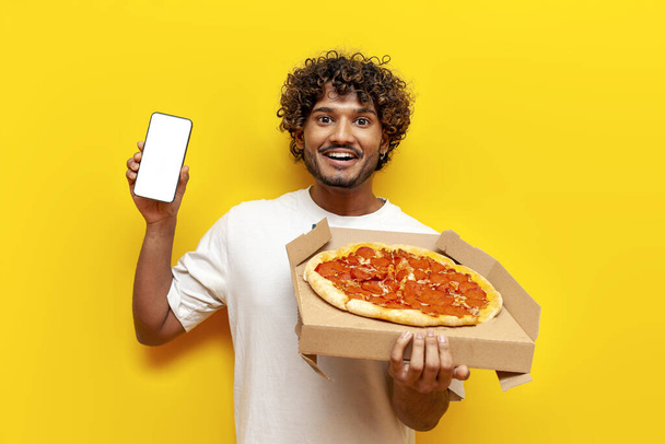 黄色い隔離された背景に空白のスマートフォン スクリーンを示すおいしいピザの箱が付いているカーリー インドの男,若者はスマートフォンでオンラインで速い食糧配達を注文します - 写真・画像
