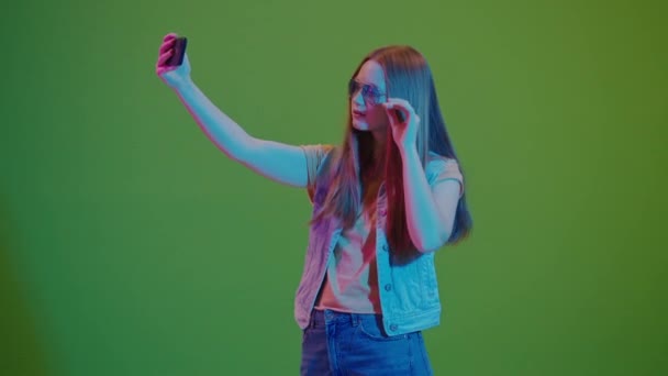 Zielony ekran. Nastolatka w neonowych światłach robi sobie selfie, promieniuje pozytywnością i duszą. Nowoczesny Snapshot Młodzieży angażującej się w kulturę cyfrową i wyrażającej swoje żywe osobowości - Materiał filmowy, wideo
