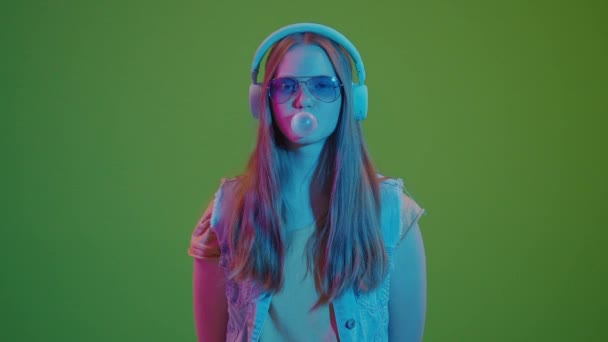 Zielony ekran. Nastolatka tańcząca w neonowych światłach, zanurzona w muzyce ze słuchawek, żując dużą różową gumę do żucia. Fuzja retro wibracji ze współczesną kulturą młodzieży i pasją do muzyki. - Materiał filmowy, wideo