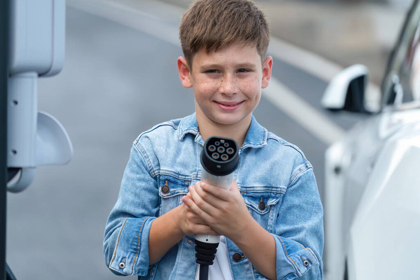 Çevre dostu EV arabalı ve EV şarj aletini kameraya doğrultmuş küçük bir çocukla yolculuk. Elektrikli araç ve şarj istasyonu çevre koruması için temiz ve sürdürülebilir enerji kullanıyor. Kalıcı - Fotoğraf, Görsel